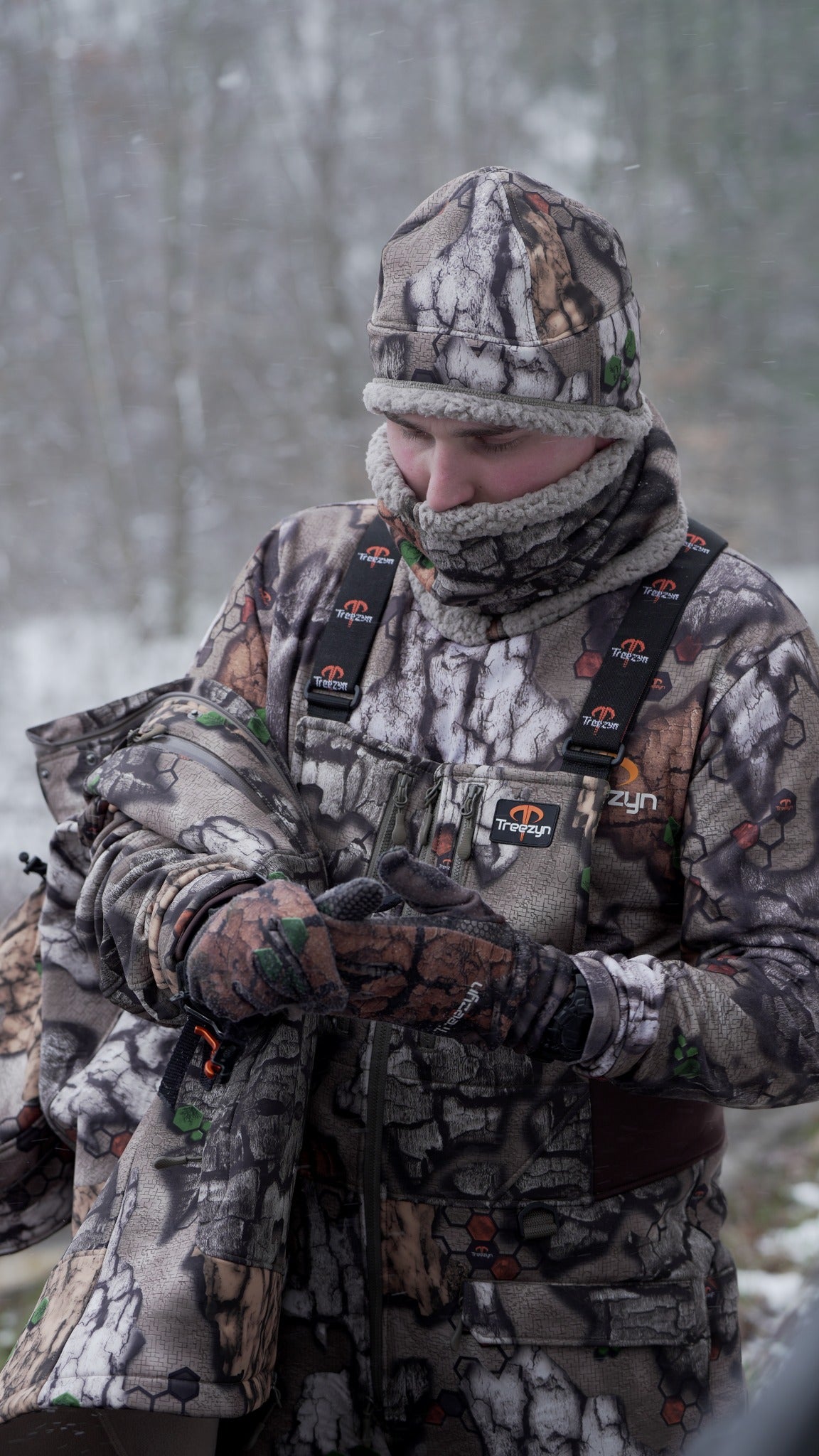 Hunter wearing Impizi Bibs with the chest zipper and branded suspenders visible, holding the removed parka